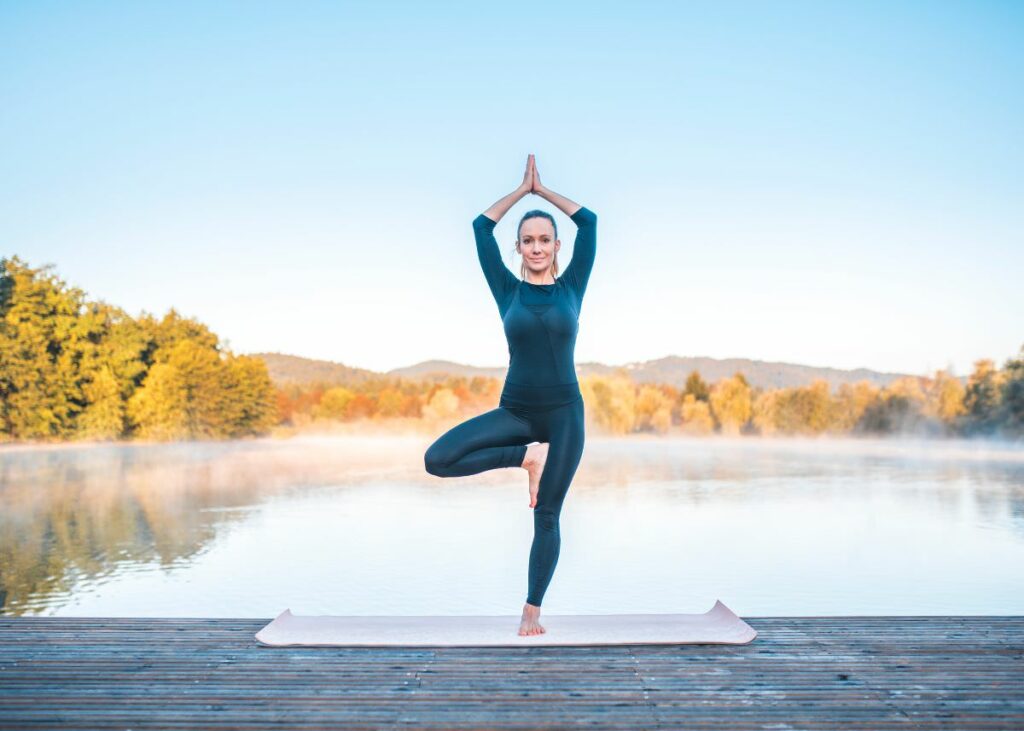 Blonde woman in Tree pose, practicing yoga on the dock of a lake.